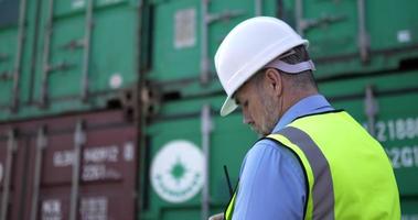 Handheld medium shot, Side view of middle-aged Caucasian Business engineer man wearing helmet checking transport container and writing on clipboard at storage port terminal video
