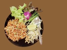 Closeup traditional Thai food Pad-Thai on black color ceramic plate with vegetable side disc isolate on brown background photo