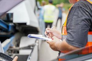 Closeup and crop insurance agent writing on clipboard while examining car after accident claim being assessed and processed on blurred damaged car truck slides and group of people background. photo