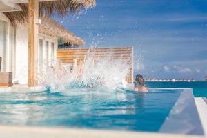 Couple having fun at the swimming pool. Man jumping into infinity pool in luxury resort with water splash over woman face and hair. Funny photo, summer honeymoon, fun outdoors activity photo