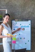 Asian businesswoman presenting business data with a board with charts, graphs hand holding a yellow notebook, And business documents lying on the table in the office, business concept. photo