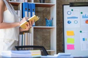 Asian businesswoman presenting business data with a board with charts, graphs hand holding a yellow notebook, And business documents lying on the table in the office, business concept. photo