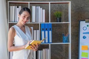 Asian businesswoman presenting business data with a board with charts, graphs hand holding a yellow notebook, And business documents lying on the table in the office, business concept. photo