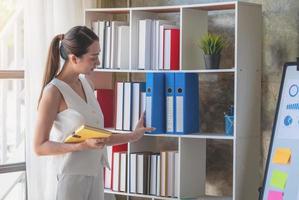 Asian businesswoman presenting business data with a board with charts, graphs hand holding a yellow notebook, And business documents lying on the table in the office, business concept. photo