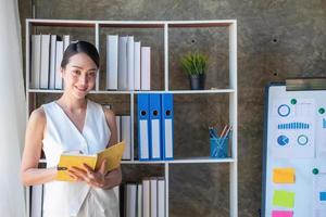 Asian businesswoman presenting business data with a board with charts, graphs hand holding a yellow notebook, And business documents lying on the table in the office, business concept. photo