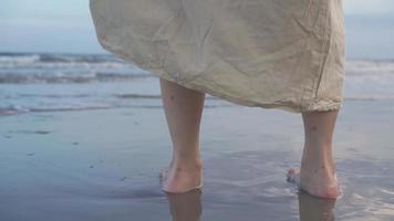 pieds de femme au ralenti marchant pieds nus de la plage à la mer. touriste en vacances d'été. femme en belle robe ondulant au coucher du soleil
