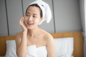 Happy young Asian woman applying face lotions while wearing a towel and touching her face. Daily makeup and skincare photo