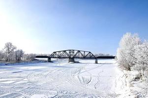 a railway bridge over a frozen river with frost covered trees on the shore photo