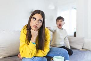 Kid anoying and disturbing exhausted and irritated mother in casual clothes holding hands on head while working at home photo