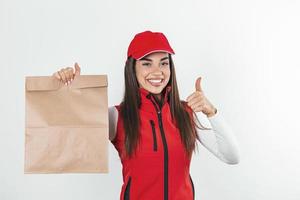 mujer de entrega en uniforme rojo sostiene un paquete de papel artesanal con comida aislada en fondo blanco, retrato de estudio. empleada con estampado de camiseta con gorra trabajando como mensajera. concepto de servicio foto