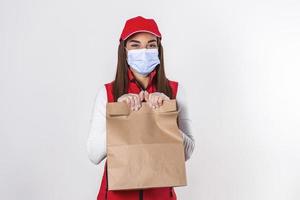 Delivery woman in red uniform hold craft paper packet with food isolated on white background, wearing medical mask and gloves. Female employee i working as courier. Service concept. photo