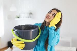 una mujer sorprendida mira el techo mientras recoge agua que se filtra en la sala de estar de su casa. mujer preocupada sosteniendo un balde mientras las gotitas de agua se filtran del techo en la sala de estar foto