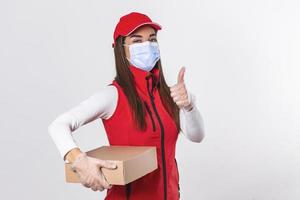 una empleada de entrega con gorra roja, camiseta en blanco, uniforme, máscara facial, guantes, caja de cartón que muestra los pulgares hacia arriba, aislada en fondo blanco. cuarentena pandemia coronavirus virus 2019-ncov concepto foto