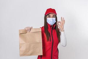 una repartidora con uniforme rojo sostiene un paquete de papel artesanal con comida aislada de fondo blanco, con mascarilla médica y guantes. empleada que trabajo como mensajera. concepto de servicio foto