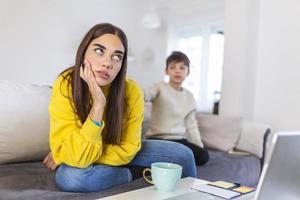 Kid anoying and disturbing exhausted and irritated mother in casual clothes holding hands on head while working at home photo