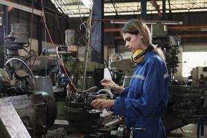 una trabajadora industrial con uniforme de protección y seguridad y casco, una joven ingeniera caucásica trabaja con una máquina metalúrgica en una fábrica de fabricación. producción profesional ocupación mecánica. foto