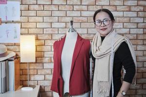 Asian mature female fashion designer stands near dressing puppet, looking at camera with smile in studio, proudly works with clothing design collection, professional boutique tailor SME entrepreneur. photo