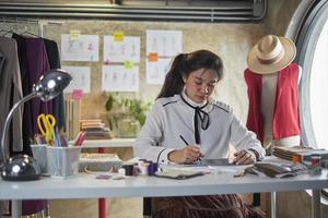 Asian young teenage female fashion designer works in studio by idea drawing sketches with tablet and craft fabric for a creative dress design collection, professional boutique tailor SME entrepreneur. photo