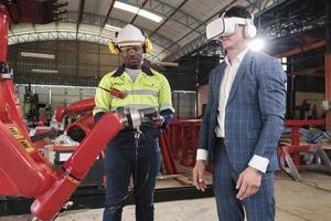 African American engineer demonstrates robotic arm control to customer with VR headsets simulator in mechanical manufacture factory, intelligent industrial machines, and futuristic technology.
