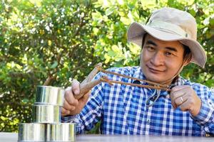 Asian man holds slingshot to play  shooting game, aim to shoot the  cans in forest. Concept, recreation activity, Hand-powered projectile weapon. Check the accuracy of target shooting. Fun game. photo