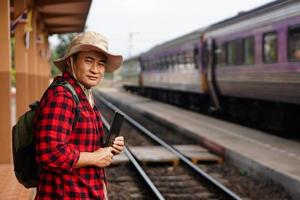 Asian man traveller is at railway station, wears hat, holds smart tablet. Concept, travel by train  in Thailand can book or by ticket online. Technology and transportation. photo