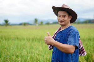 Portrait of Asian man farmer wears hat, blue shirt, thumbs up, Feels confident, stands at paddy field. Concept, Agriculture occupation. Thai farmers grow  organic rice. Copy space for adding text .