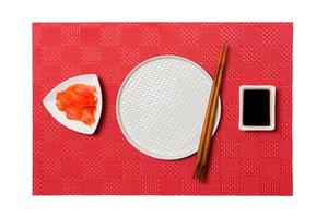 Empty round white plate with chopsticks for sushi and soy sauce, ginger on red mat sushi background. Top view with copy space for you design photo