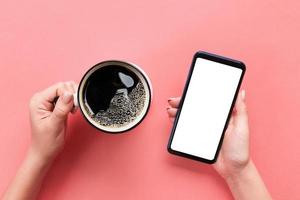 Female hands holding black mobile phone with blank white screen and mug of coffee. Mockup image with copy space. Top view on pink background, flat lay photo