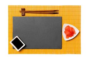 Empty rectangular black slate plate with chopsticks for sushi, ginger and soy sauce on yellow bamboo mat background. Top view with copy space for you design photo