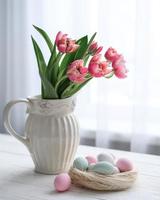 A nest with eggs in soft pastel colors and a vase with tulips on a table near the window photo