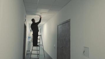 en bemästra elektriker är stående på en stege och ändring en ljus Glödlampa i en mörk korridor video