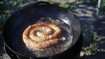 salsicha caseira deliciosa frita em uma churrasqueira. um anel de salsicha é frito em óleo fervente em uma frigideira redonda ao ar livre. video