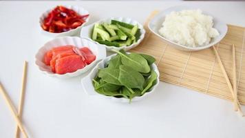 Ingredienser för framställning sushi i skålar på en vit tabell bakgrund. lax fisk, ris, peppar, sallad och gurka närbild. ätpinnar för sushi och en bambu servett för framställning rullar. video