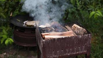 brandhout is brandend in de rooster in buitenshuis. aanmaakhout brandhout Aan een zomer avond voor barbecue. hout logboeken zijn roken in de metaal koperslager dichtbij omhoog. wit dik rook stijgt van de brander. video