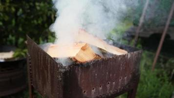 Wood logs are smoking in the metal brazier close up. A man throws firewood into a smoking grill in his backyard. White thick smoke rises from the roaster. Firewood is burning in the grill in outdoor.