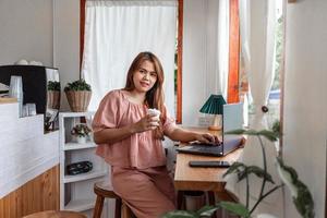 A happy female at a cafe using a laptop in hand and a paper cup of coffee looking at the camera. young white woman with long hair sitting in a coffee shop busy working on her laptop. photo