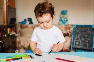 un niño pequeño dibuja en hojas de papel sobre la mesa con lápices de colores foto