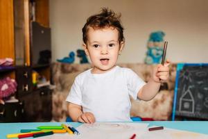 un niño pequeño dibuja en hojas de papel sobre la mesa con lápices de colores foto