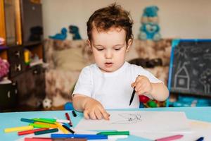 un niño pequeño dibuja en hojas de papel sobre la mesa con lápices de colores foto