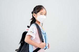 niña asiática lleva una mochila con máscara de respirador para proteger el brote de coronavirus y apunta con la mano al fondo en blanco, nuevo virus covid-19 foto