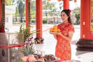 feliz Año Nuevo Chino. hermosa joven con vestido tradicional cheongsam qipao sosteniendo naranjas frescas en el templo budista chino. orar por el mejor deseo bendición y buena suerte foto