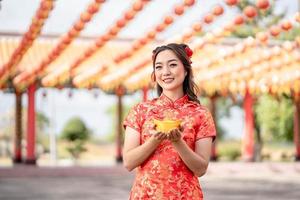 una joven asiática con un vestido tradicional cheongsam qipao sosteniendo dinero de oro antiguo en un templo budista chino. celebrar el año nuevo lunar chino, vacaciones de temporada festiva. emoción sonrisa foto
