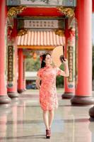 Happy Chinese new year. Beautiful lady wearing traditional cheongsam qipao dress holding fan while visiting the Chinese Buddhist temple. Emotion smile