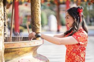 feliz Año Nuevo Chino. mujer asiática con vestido tradicional cheongsam qipao poniendo varitas de incienso para la mejor bendición y buena suerte en el templo budista chino. foto