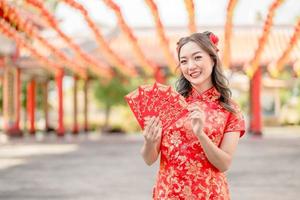 Portrait Beautiful Asian woman wearing traditional cheongsam qipao costume holding ang pao, red envelopes in Chinese Buddhist temple. Chinese text means great luck great profit. photo