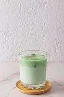 A wide glass of natural green tea matcha latte stands on a wooden plate and a marble white table. health-improving, invigorating agent. photo