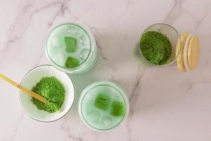 vista superior de dos vasos con café con leche matcha frío y cubitos de hielo verde del partido sobre un fondo blanco de mármol. polvo de té verde en un bol y tarro.