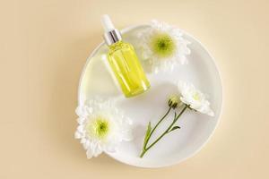 A bottle with a dropper and a white cap lies on a white ceramic plate and delicate flowers. top view. beige background. natural cosmetics. photo