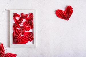 corazones diy de papel rojo en un marco de fotos vacío y hojas sobre un fondo concreto.