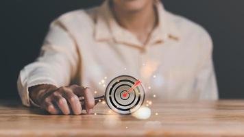 magnifying glass in businessman hand and arrow icon hitting center of dartboard target,Setting business goals and focused concepts, Organizational growth and objectives, marketing strategy photo